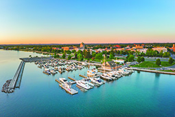Aerial view of a marina filled with boats and yachts on calm blue water, adjacent to a waterfront cityscape at sunset. The shoreline is lined with green trees, open spaces, and buildings, with a soft gradient sky transitioning from blue to orange in the background.Aerial view of a marina filled with boats and yachts on calm blue water, adjacent to a waterfront cityscape at sunset. The shoreline is lined with green trees, open spaces, and buildings, with a soft gradient sky transitioning from blue to orange in the background.