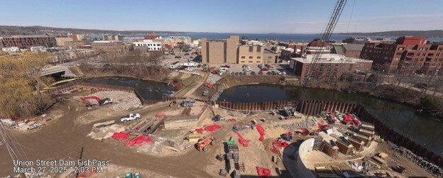 Aerial view of the Union Street Dam FishPass construction site taken on March 27, 2025, at 12:03 PM. The site is active with numerous construction vehicles and equipment spread throughout, including excavators and dump trucks. Bright red coverings and materials are visible across multiple work zones. Sheet piling remains in place, dividing parts of the river. The surrounding city buildings, roads, and distant view of the bay under a mostly clear sky are also visible.
