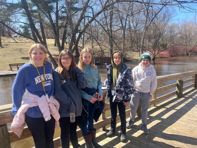 Five middle school students stand on a wooden boardwalk near a calm river, smiling at the camera. The students are dressed in casual, warm clothing including hoodies and jackets, with leafless trees and a grassy park visible in the background on a sunny spring day.